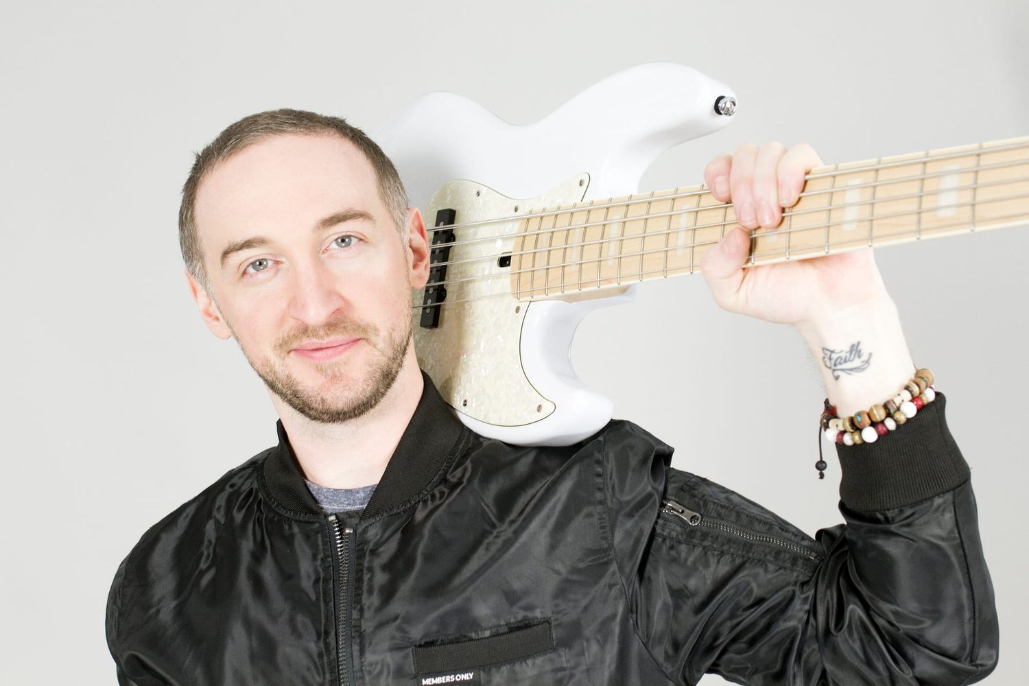 Alex in the studio in 2018, in a black bomber jacket, smiling, a white five-string bass resting on his shoulder.