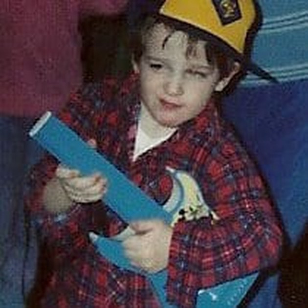 A faded childhood snapshot from the family collection: Alex in a plaid shirt and cap, holding a toy guitar.