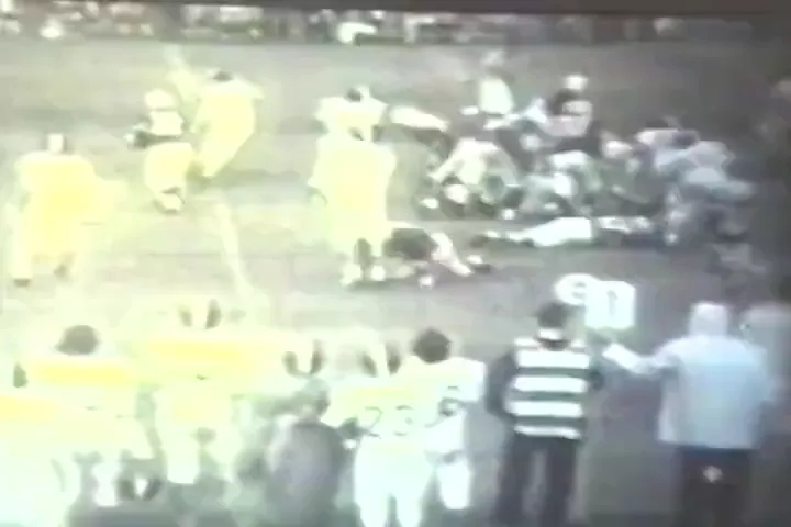 A frame of 8mm film from 1971: a high school football game, players in light jerseys on a grey field, a crowd behind.