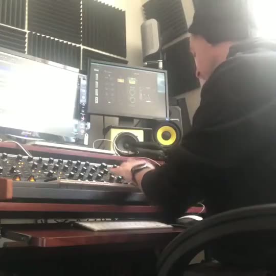 A home studio: a synthesizer with rows of knobs in the foreground, a screen and speakers behind it, Alex at the right edge.